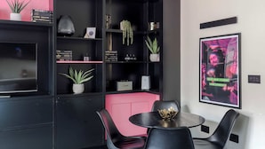 Un interno moderno caratterizzato da una combinazione di colori nero e rosa, con una libreria nera piena di libri e piante, un tavolo rotondo con sedie e un poster appeso al muro.