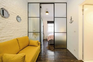 A room with a yellow sofa, a sliding door with frosted panels, a bed with a pink blanket, and a small window. - The Cheeky Hideaway - Borough - by Frankie Says (London)