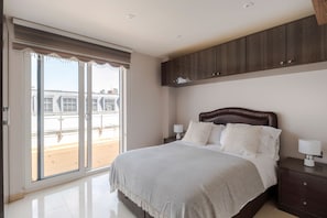 Un dormitorio con un gran ventanal corredero que da a un balcón, una cama con cabecera, dos mesitas de noche con lámparas y un armario de madera sobre la cama.
