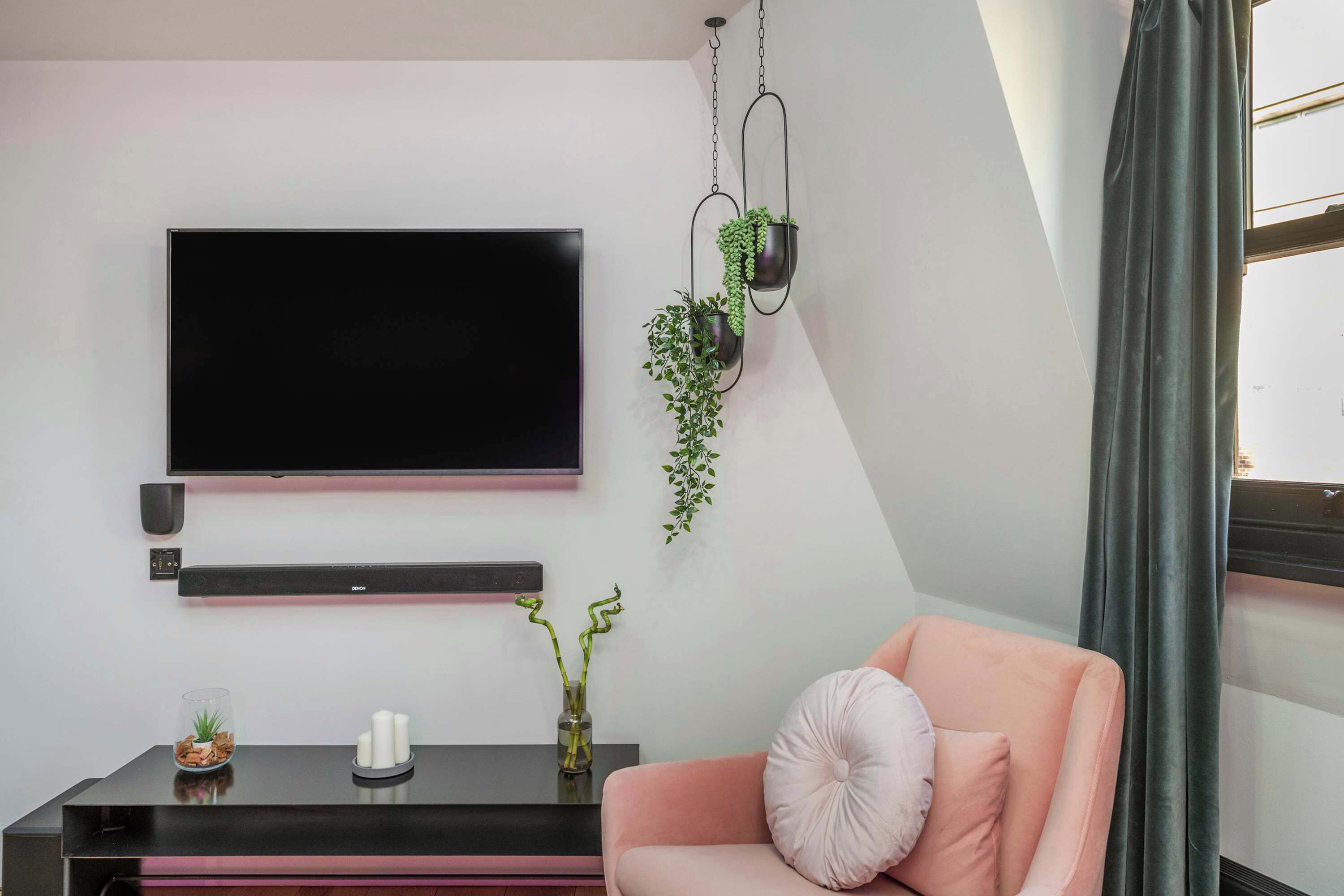 A modern living room with a flat-screen TV, a black console table, a pink sofa, and hanging plants.
