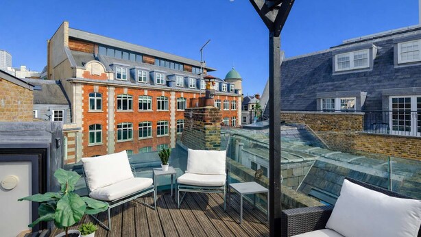 A rooftop terrace with glass railings, white cushioned seating, and a potted plant. - The Acre Penthouse - Covent Garden - by Frankie (London)