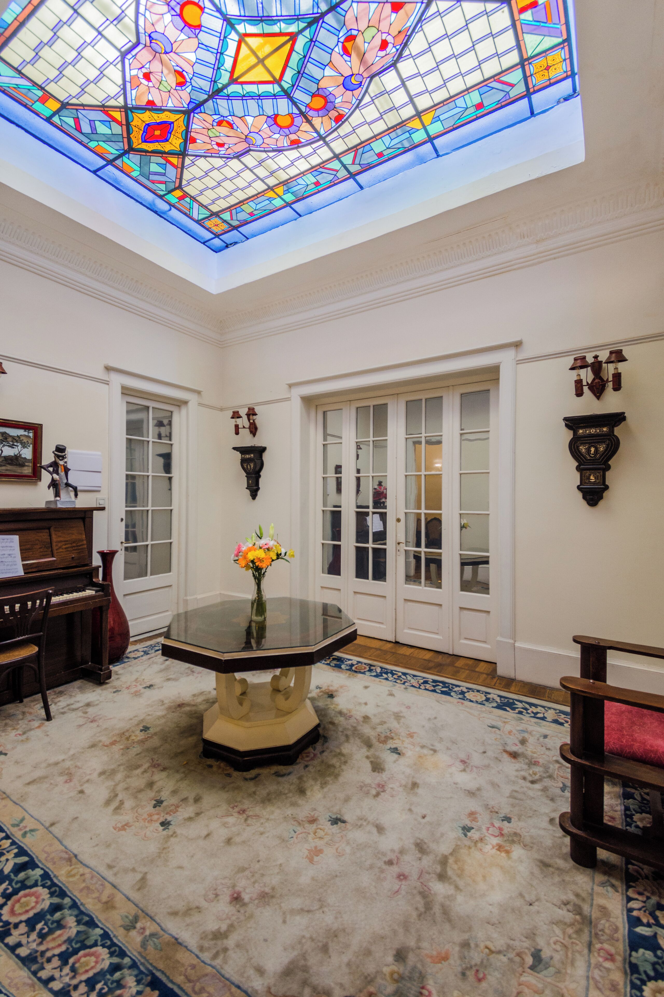 Una habitación con techo de vitrales, un piano, una mesa adornada con flores y una alfombra estampada.