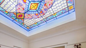 Una stanza con soffitto in vetro colorato, un pianoforte, un tavolo con fiori e un tappeto decorato.