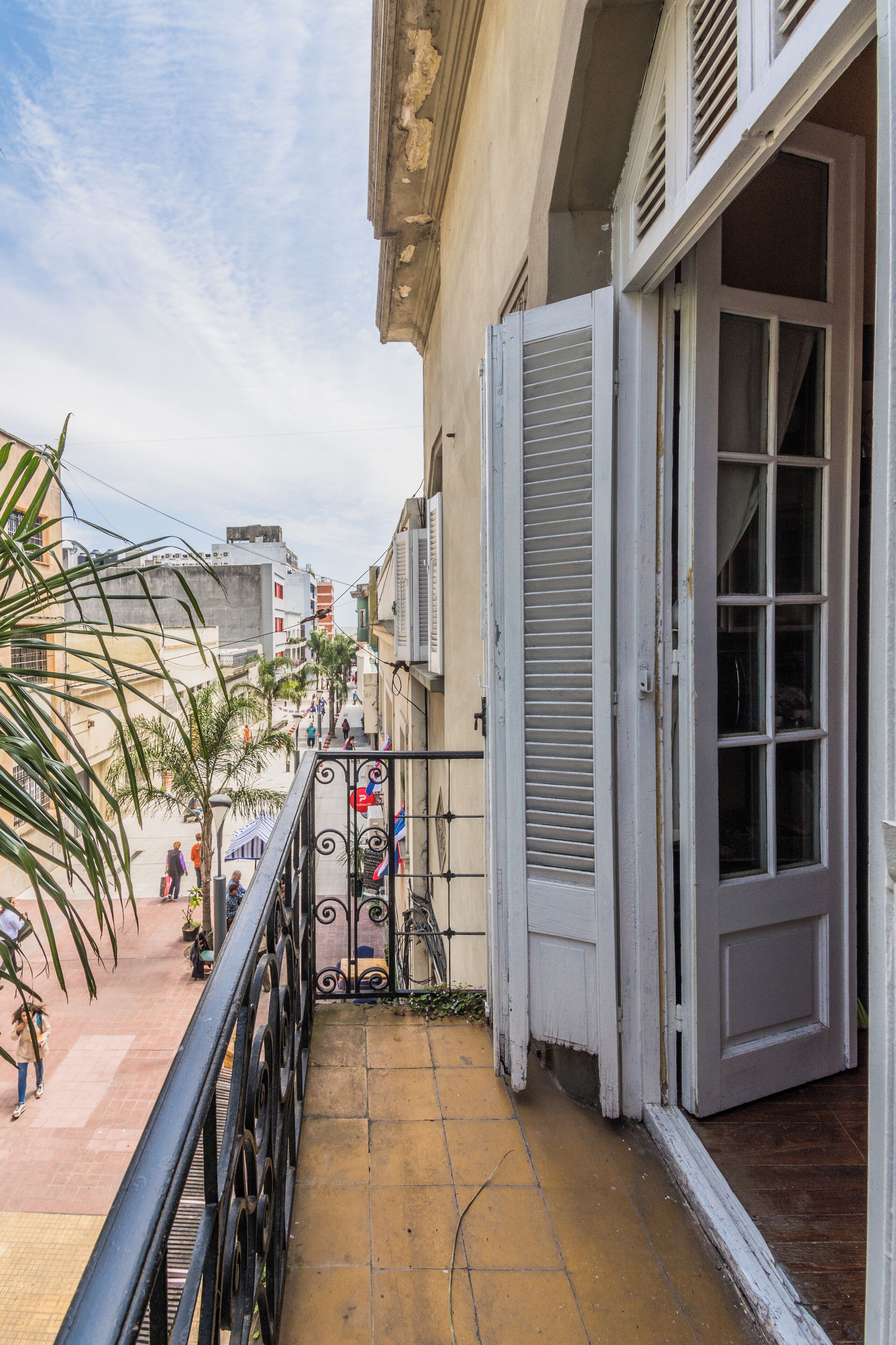 A narrow outdoor corridor with a metal railing, a door with shutters, and a tiled floor.