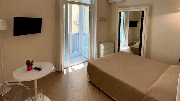Camera d'albergo con un letto, un tavolino con un cactus, una sedia, una TV a muro e un'ampia finestra con tende.
