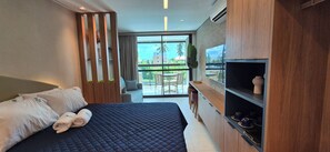 Ein modernes Hotelzimmer mit einem großen Bett, einem Fernseher, einem Balkon mit Blick auf Palmen und einem Holzkasten.