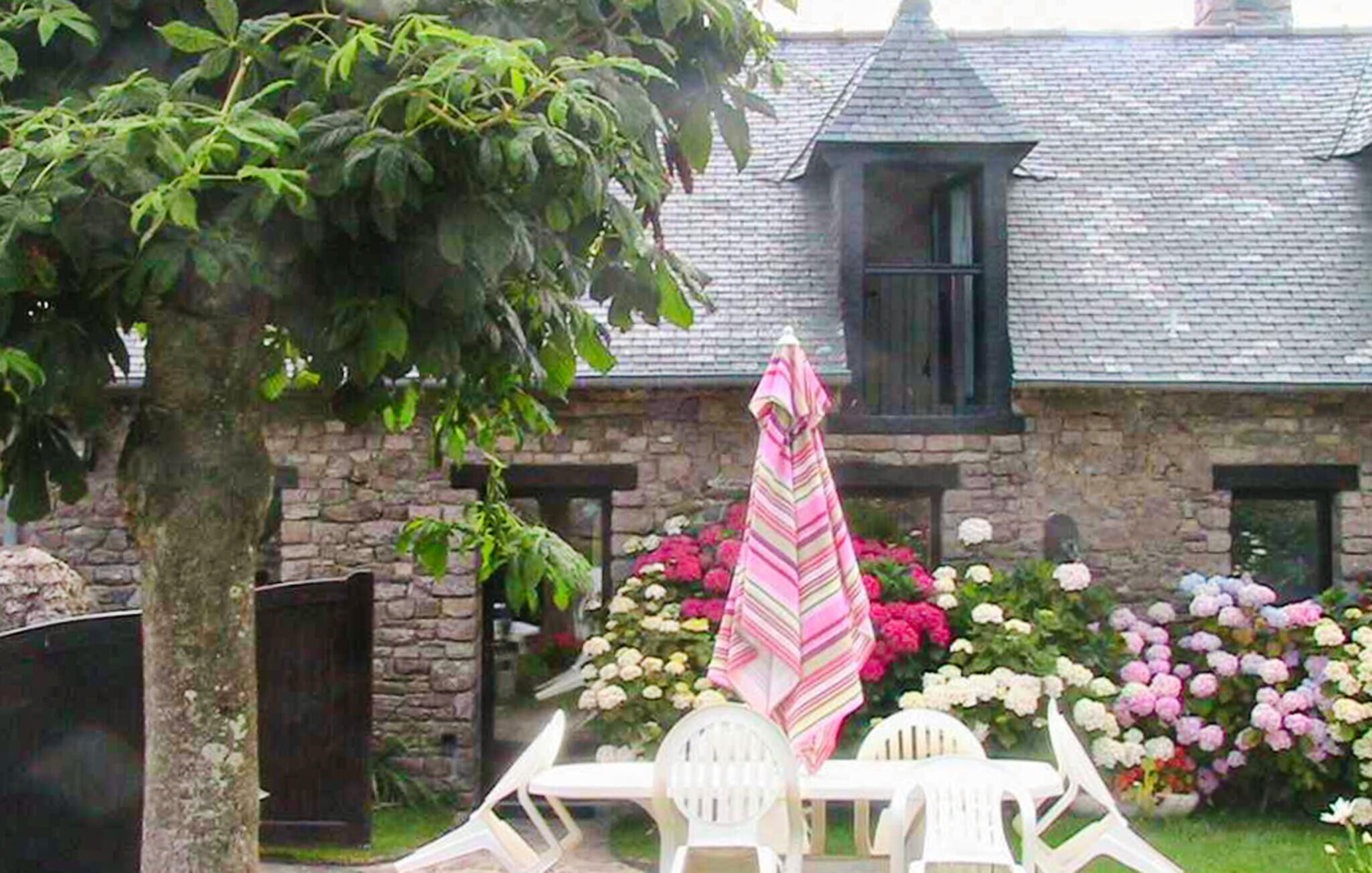 A stone building with a pitched roof, a window, and a door. There is a tree in the foreground and a table with chairs and a striped umbrella in the background.
