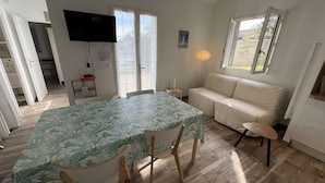 A room with a dining table, chairs, a sofa, a TV, and a window. - 148 – Charming House with Large Garden – Ideal for 4 to 6 Guests ?? (SOULAC SUR MER)