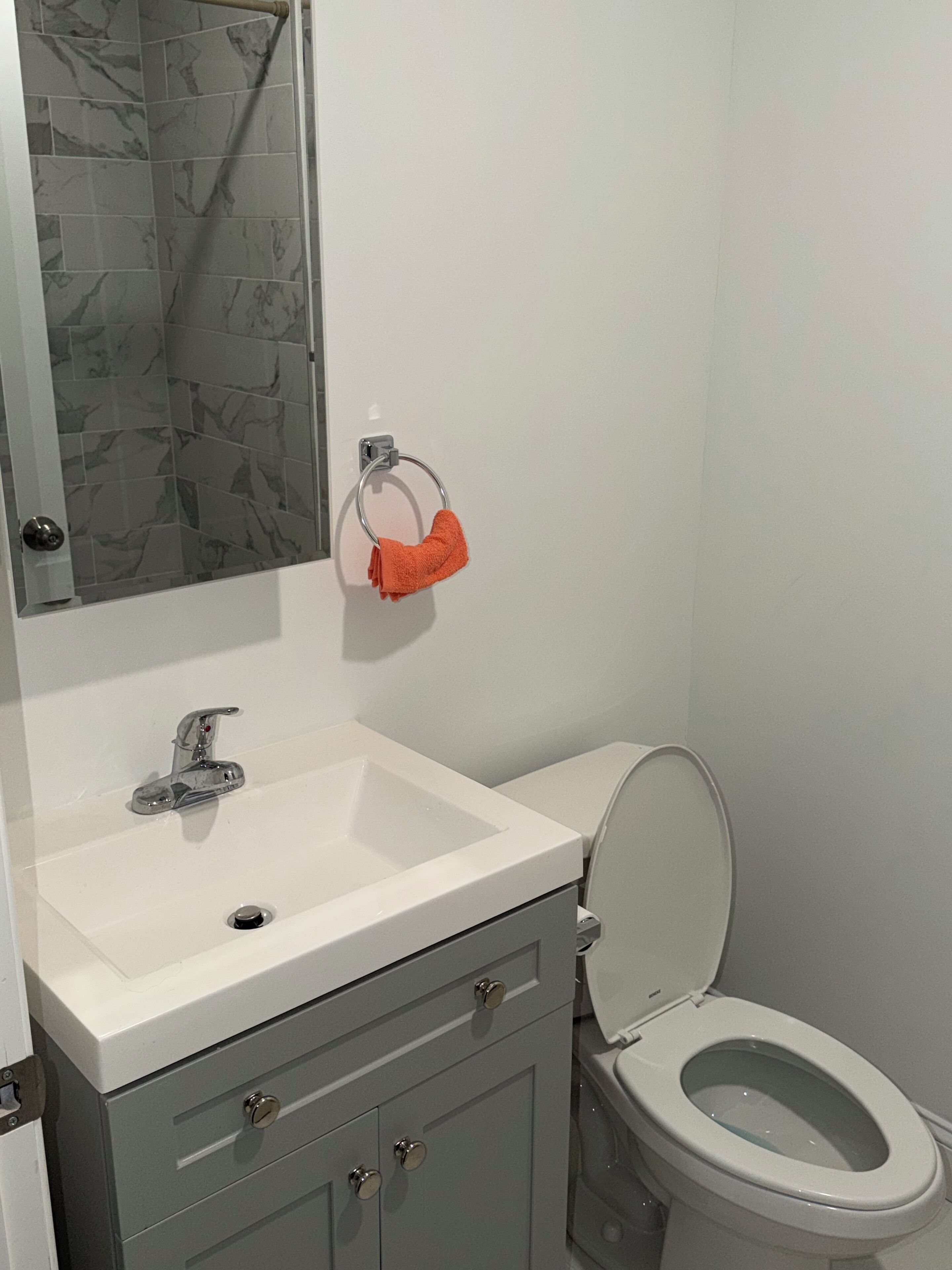 Un baño con lavamanos blanco, un espejo, un toallero con una toalla naranja, inodoro y zona de ducha con baldosas de mármol.