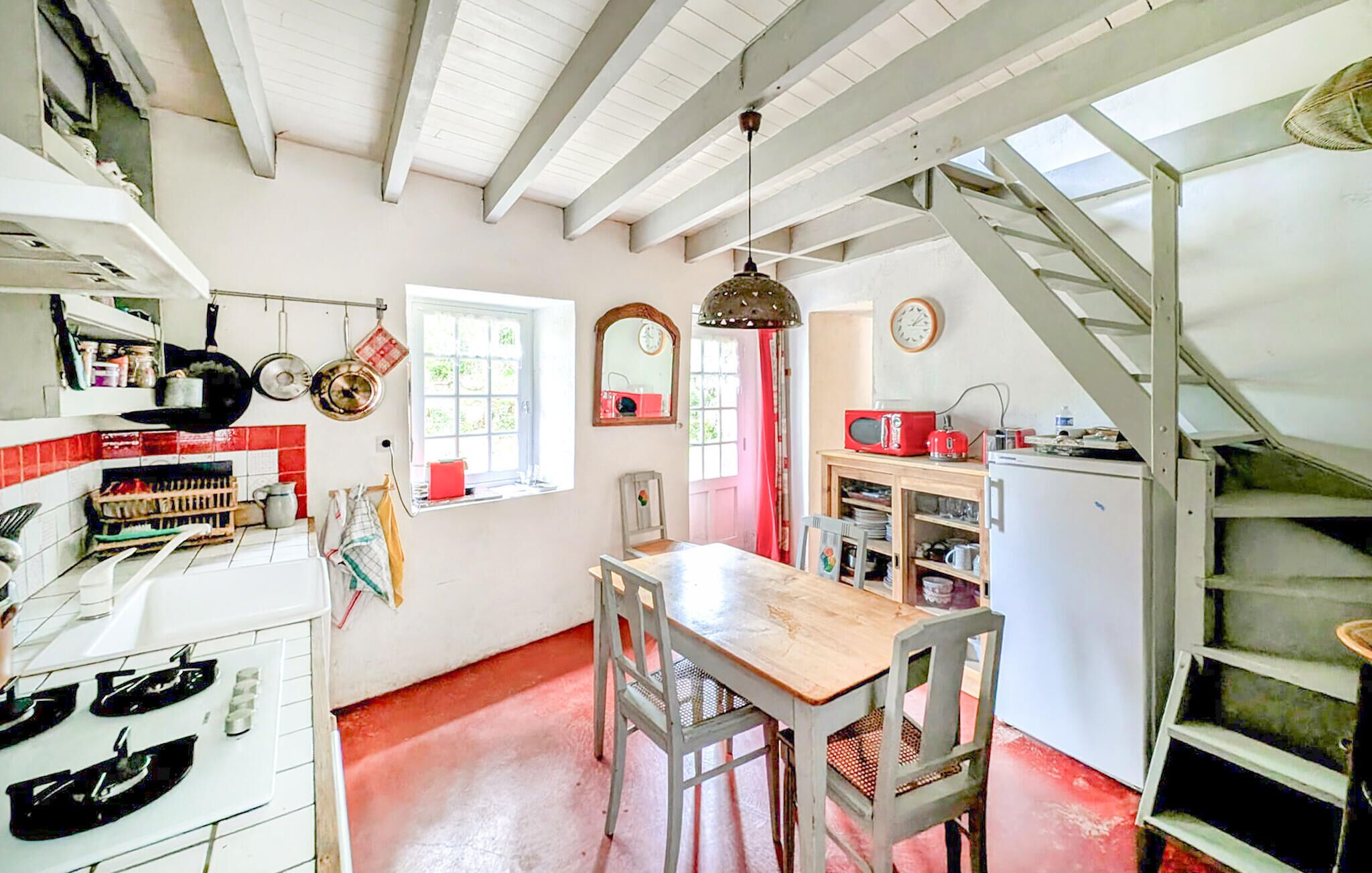 Una cucina con mobili bianchi, un tavolo di legno e una finestra con cornice rossa.