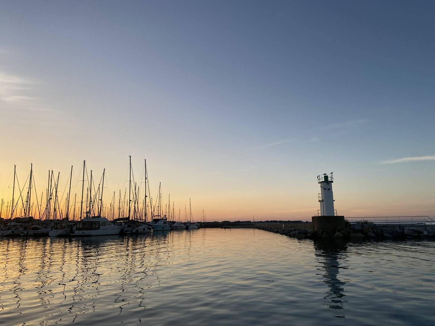 Een jachthaven met diverse zeilboten afgemeerd en een vuurtoren op de achtergrond.