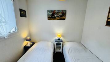 Una habitación pequeña y limpia con dos camas individuales, una ventana con cortina, un cuadro en la pared y una lámpara en el techo.