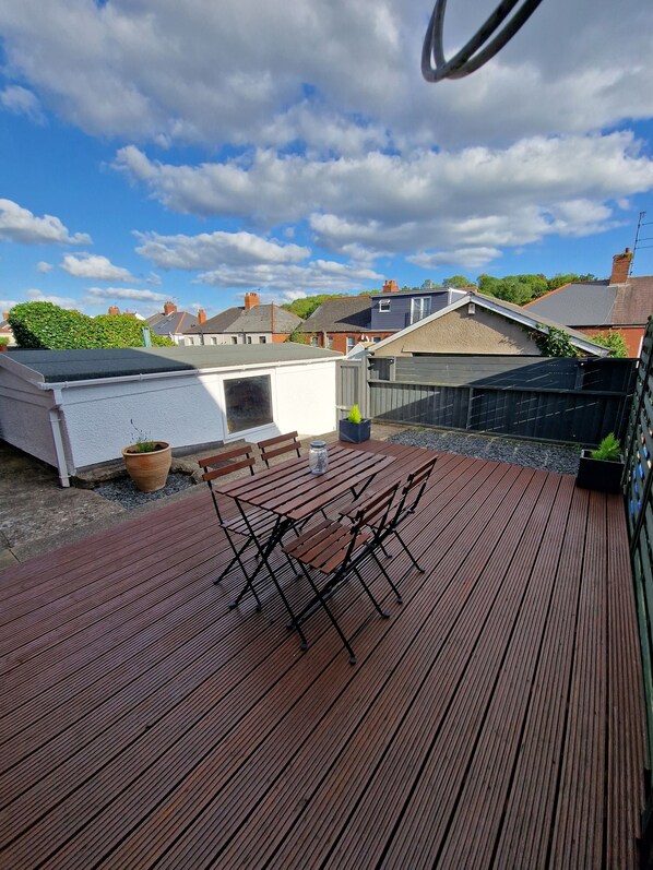 A wooden deck with a table and chairs, surrounded by a garden and houses in the background.