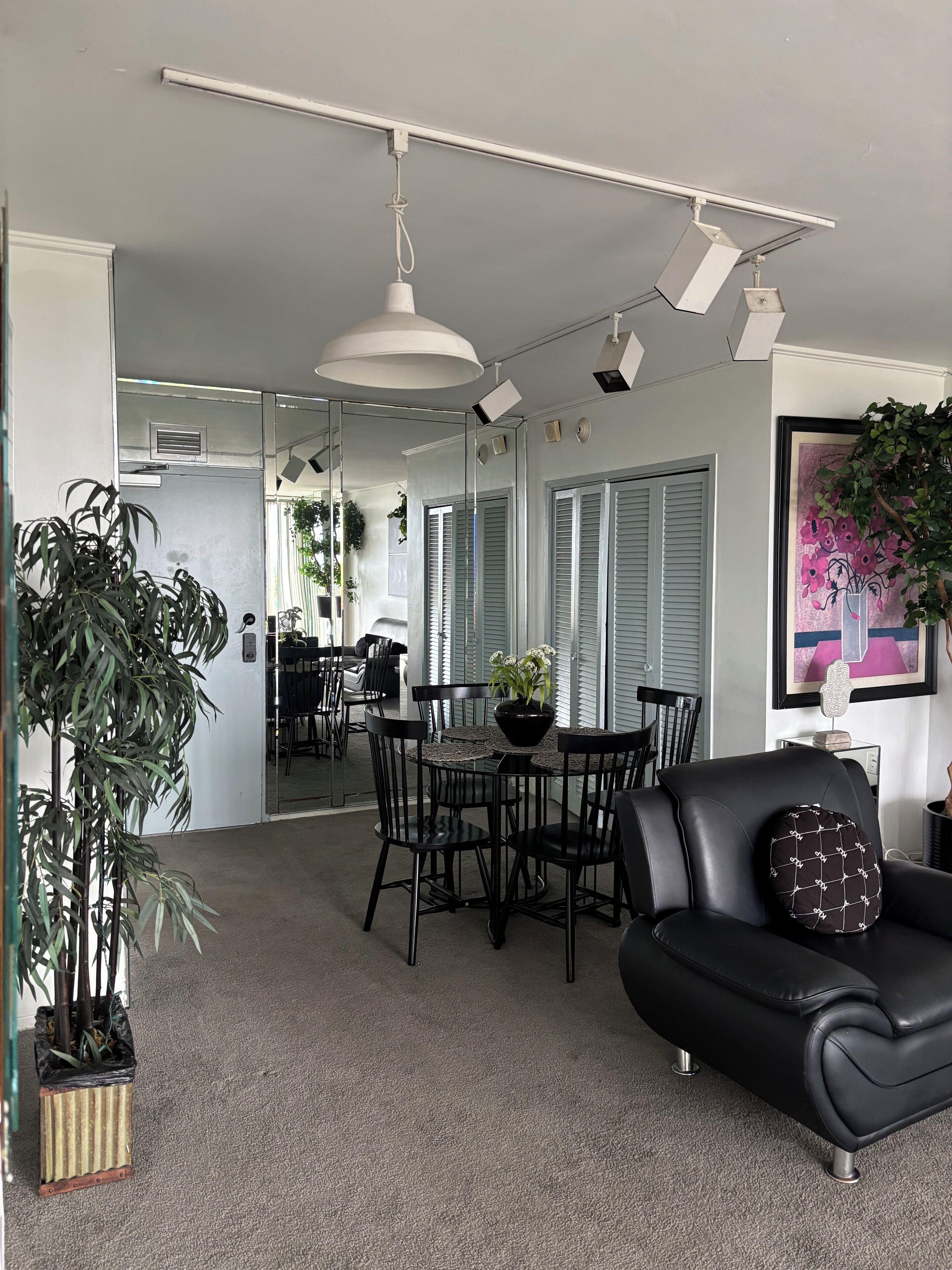 A modern living room with a black leather sofa, a dining table with chairs, and a large mirror reflecting the room. There are potted plants and a framed artwork on the walls.