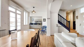 A spacious living area with a wooden dining table, blue chairs, a white sofa, and a staircase with a dark railing. - Charmante maison proche mer (Royan)
