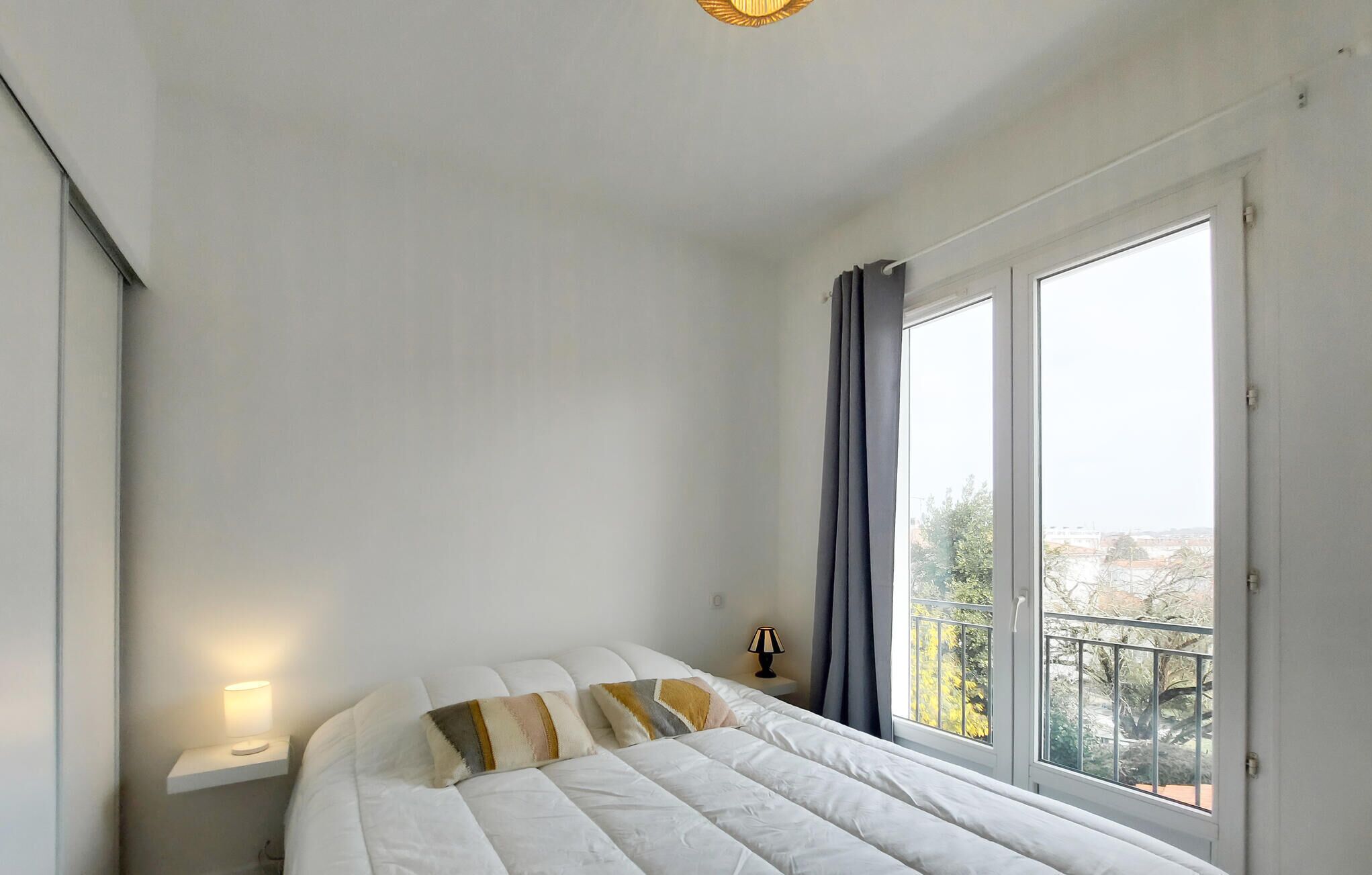 Ein Schlafzimmer mit Bett, Nachttischlampe, einem Fenster mit Vorhängen und Blick ins Freie.