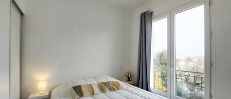 Ein Schlafzimmer mit Bett, Nachttischlampe, einem Fenster mit Vorhängen und Blick ins Freie.