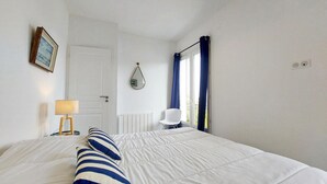 A bedroom with a large bed, a bedside table with a lamp, a chair, a window with curtains, and a door. - Charmante maison proche mer (Royan)