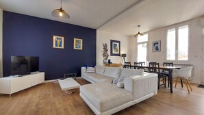 A modern living room with a white sofa, a flat-screen TV, and a dining area with a table and chairs. - Charmante maison proche mer (Royan)