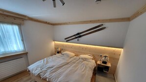 Un dormitorio con cama de madera, cabecera con porta esquíes, mesita de noche con lámpara y ventana con cortinas.