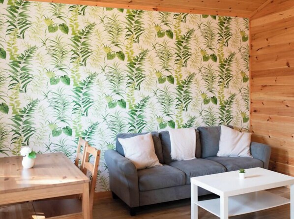 A cozy room with a grey sofa, a white coffee table, and a wooden dining table with chairs. The walls are adorned with a leafy green wallpaper.