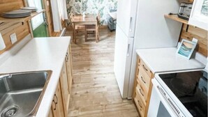 A compact kitchen with wooden cabinets, a sink, a refrigerator, and a dining table with chairs. - The Pine, Lawrencetown (West Porters Lake)
