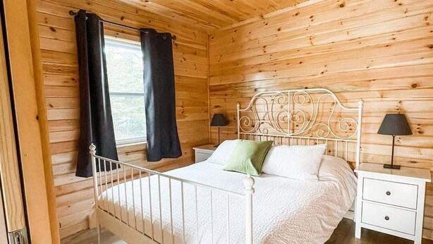 A wooden cabin bedroom with a metal bed frame, a white bedside table with a lamp, and a window with black curtains. - The Pine, Lawrencetown (West Porters Lake)