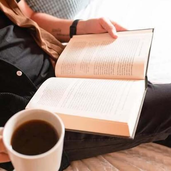 Eine Person sitzt auf einem Bett, ein Buch und eine Tasse Kaffee in der Hand.