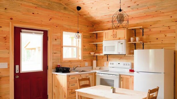 A compact kitchen with wooden walls, a red door, a white refrigerator, a microwave, a stove, and a dining table with chairs. - The Redwood Studio - Lawrencetown Lodge (West Porters Lake)
