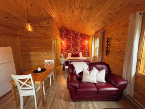 A wooden cabin interior with a dining area, a red leather sofa, a bed with floral wallpaper, and a hanging light fixture.