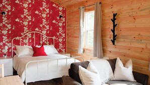 A room with a wooden ceiling and walls, a bed with white bedding and a red pillow, a chair with white cushions, and a window with curtains. - The Redwood Studio - Lawrencetown Lodge (West Porters Lake)