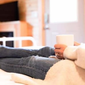 A person sitting on a bed with a white blanket, holding a white mug.