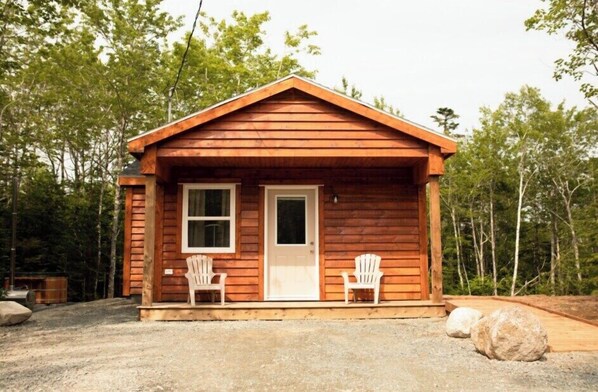 A small wooden cabin with a gabled roof, two windows, and a door. There are two Adirondack chairs outside.
