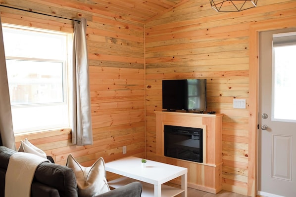 Una habitación acogedora con paredes de madera, un televisor de pantalla plana sobre un soporte de madera, una chimenea, una mesa de centro blanca y un sofá con cojines.