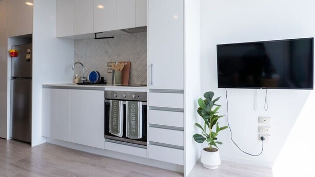 A modern kitchen with white cabinets, a stainless steel refrigerator, a built-in oven, and a flat-screen TV mounted on the wall. - Urban Sunlit Nest in the Heart of CBD (Auckland Central)