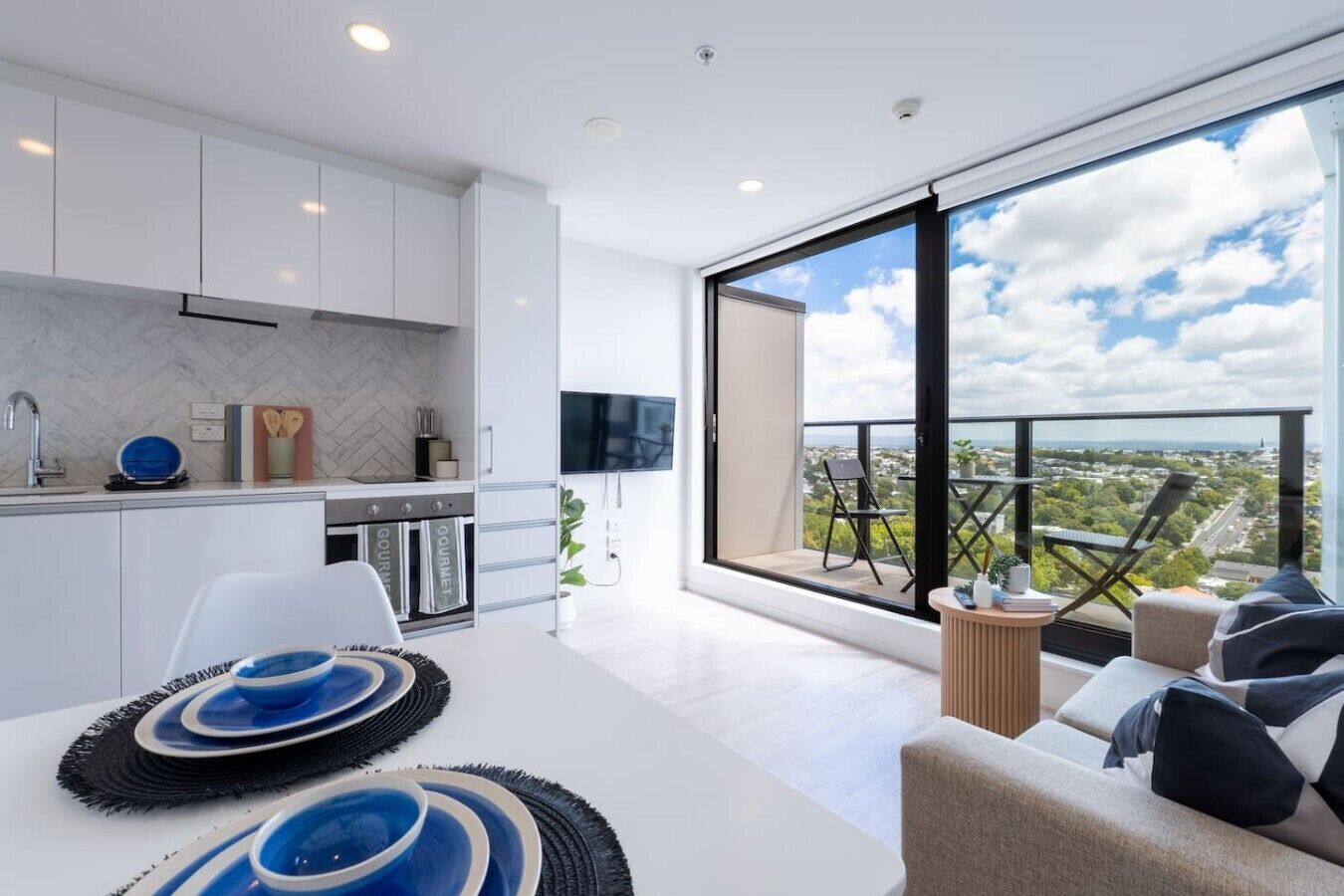 A modern kitchen with white cabinets, a dining area with a white table, and a balcony with a view of the city.