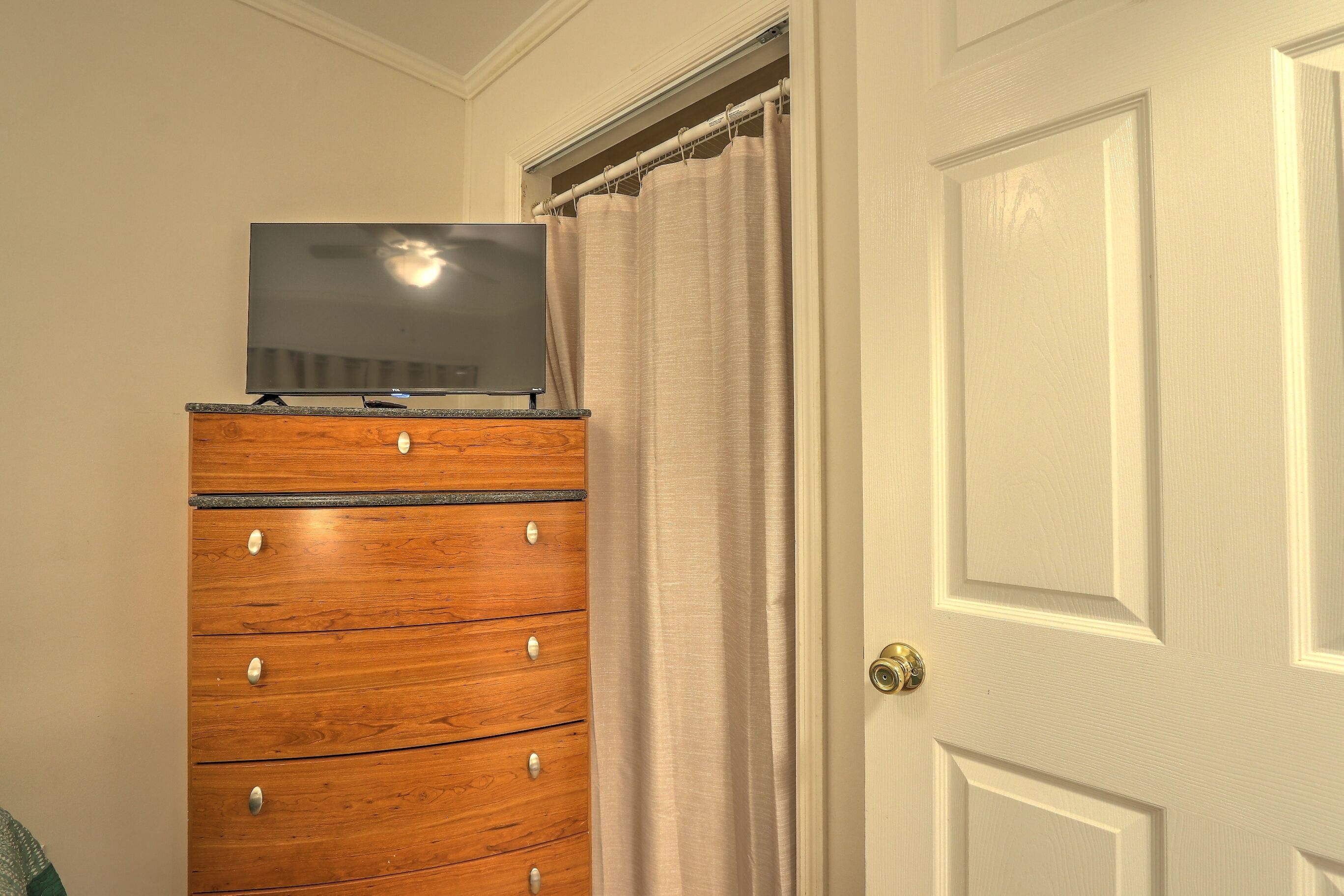 A wooden dresser with a flat-screen TV on top, a white door, and a beige curtain.