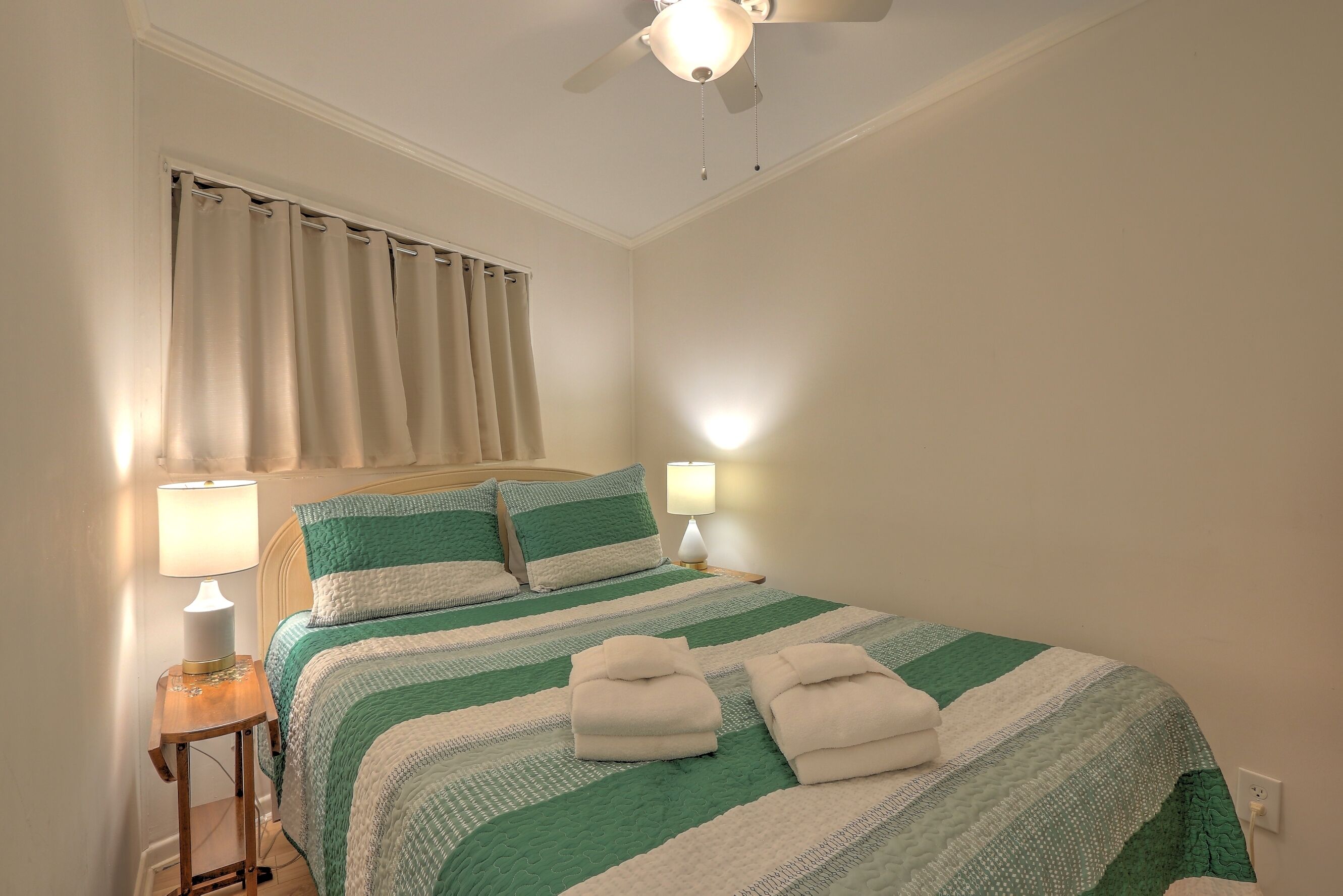 A neatly made bed with a striped green and white bedspread, two pillows, and two folded towels. There is a bedside lamp on a wooden nightstand, a ceiling fan, and a window with curtains.