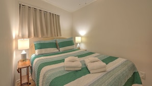 Ein ordentlich bezogenes Bett mit einer gestreiften Bettdecke in Grün und Weiß, zwei Kissen und zwei gefaltete Handtücher. Auf dem Nachttisch steht eine Nachttischlampe, es gibt einen Deckenventilator und ein Fenster mit Vorhängen.