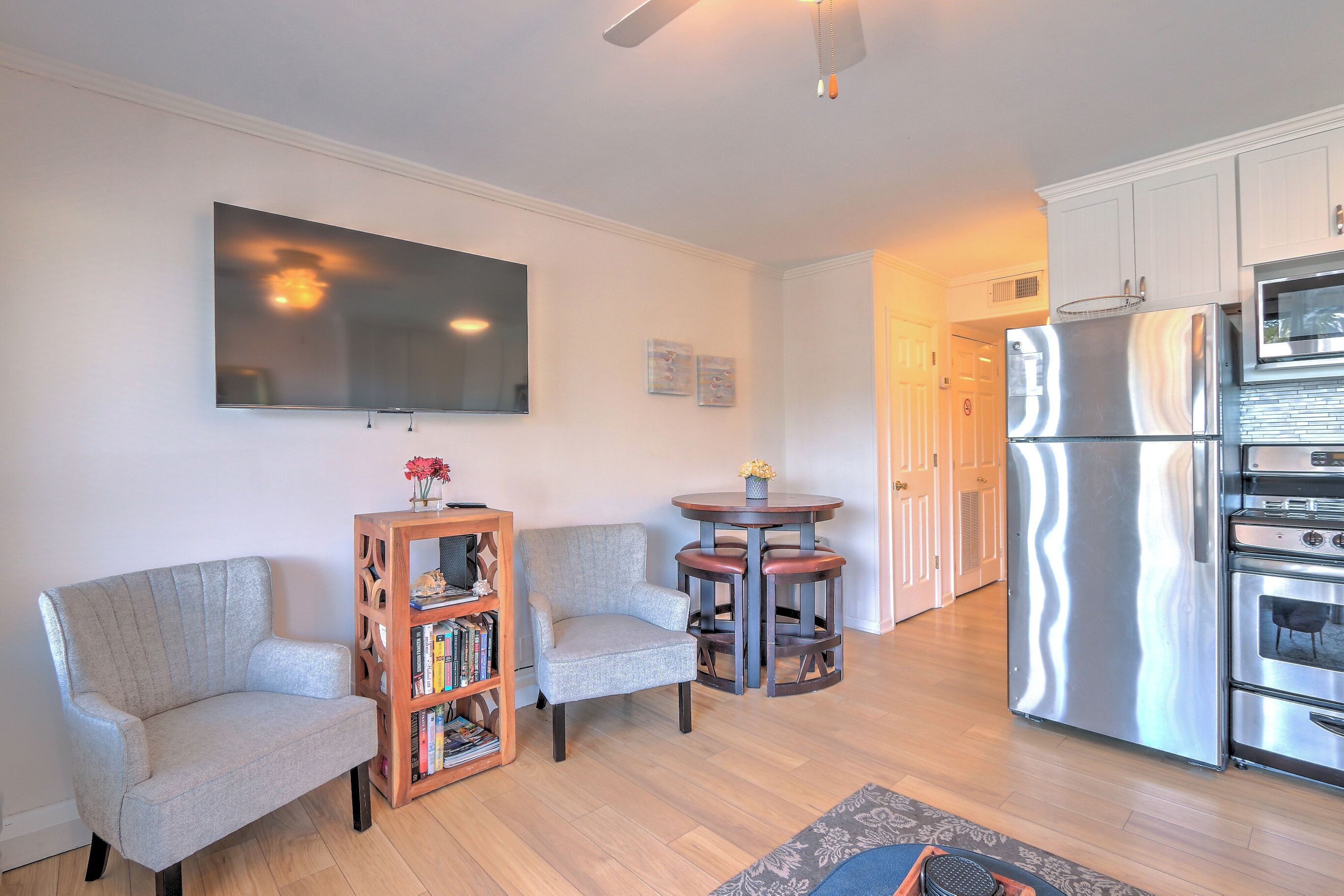 Una sala de estar con un sofá gris, una estantería de madera, una mesa redonda con sillas, un televisor de pantalla plana en la pared y un refrigerador de acero inoxidable en la zona de la cocina.