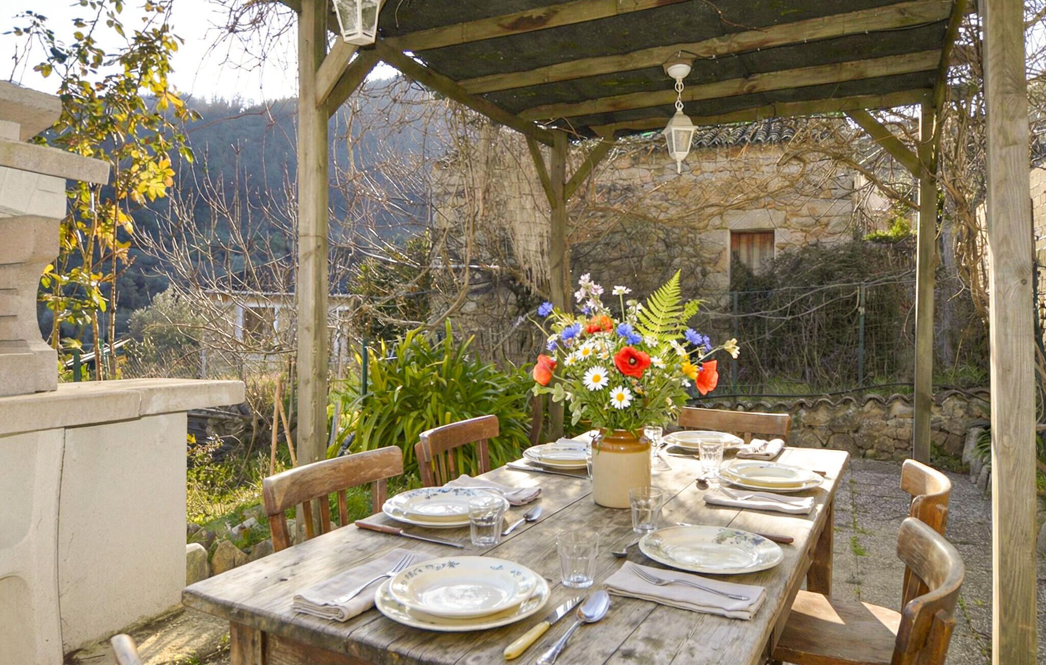 An outdoor wooden table set for dining with a flower vase, surrounded by stone walls and a stone building in the background.