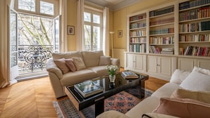 Een woonkamer met een beige bank, een bijzettafeltje met boeken, een boekenkast en een uitzicht naar buiten door grote ramen.