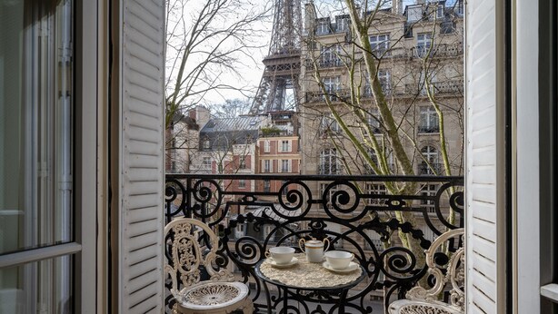 Een balkon met sierlijk ijzerwerk, twee witte stoelen en een kleine ronde tafel met een theepot en kopjes.