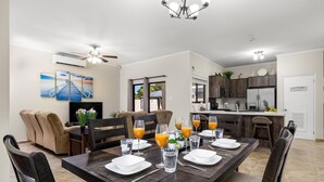 A dining area with a dark wooden table set for six, surrounded by chairs. There is a television on the wall, a ceiling fan, and a kitchen area visible in the background. - Tebenki Retreat - Entire House (Boegoeroei)