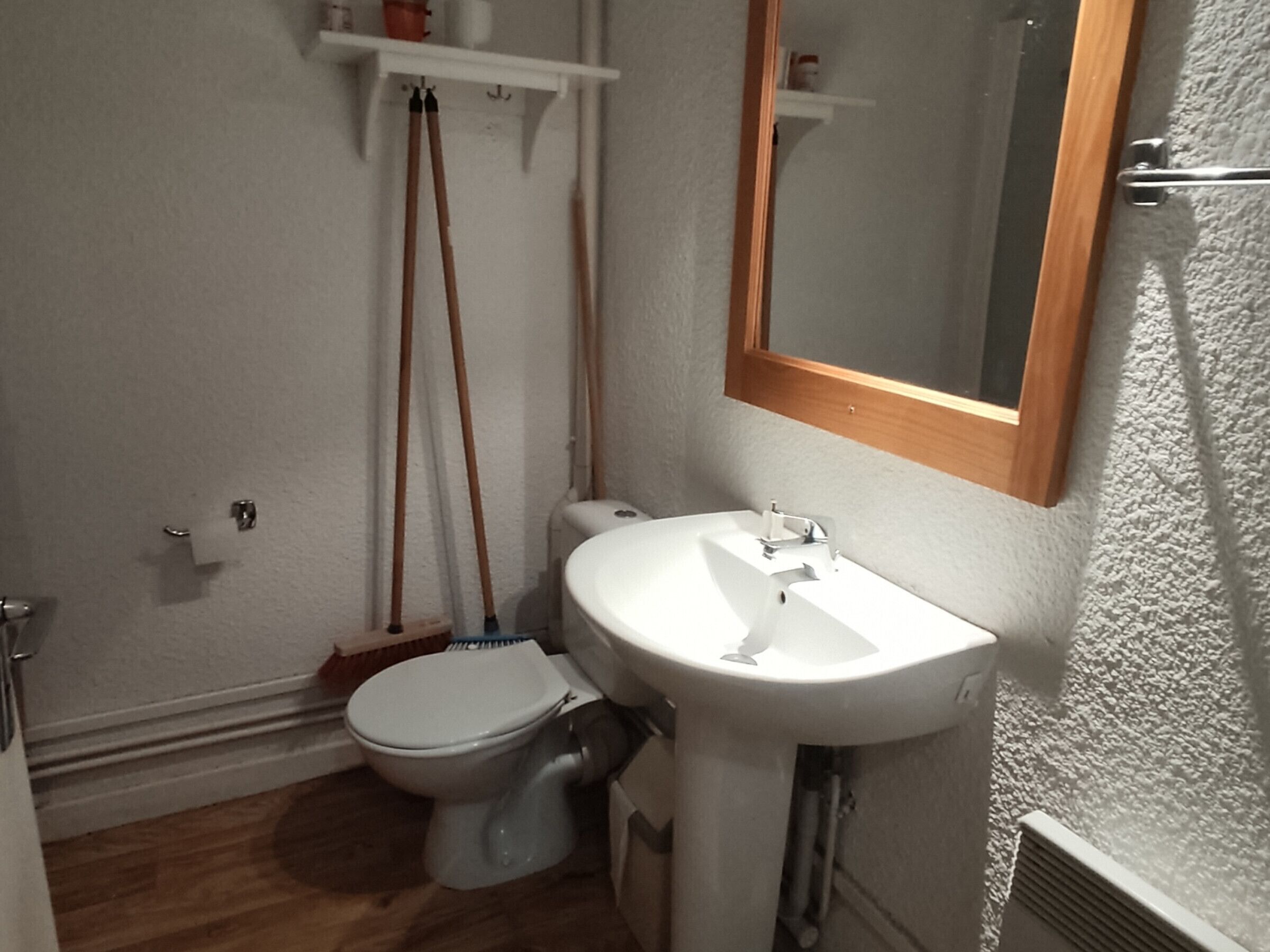 Een kleine badkamer met toilet, wastafel en spiegel. Er staan schoonmaakmiddelen aan de muur en een plank boven het toilet.