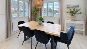 Un comedor con una mesa de madera, sillas negras, una lámpara colgante, una planta en maceta y vistas al exterior a través de ventanas amplias.