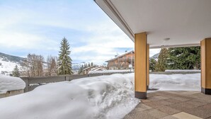 Ett takförsett utomhusområde med snö på marken, utsikt över byggnader och berg i bakgrunden samt ett klinkergolv.