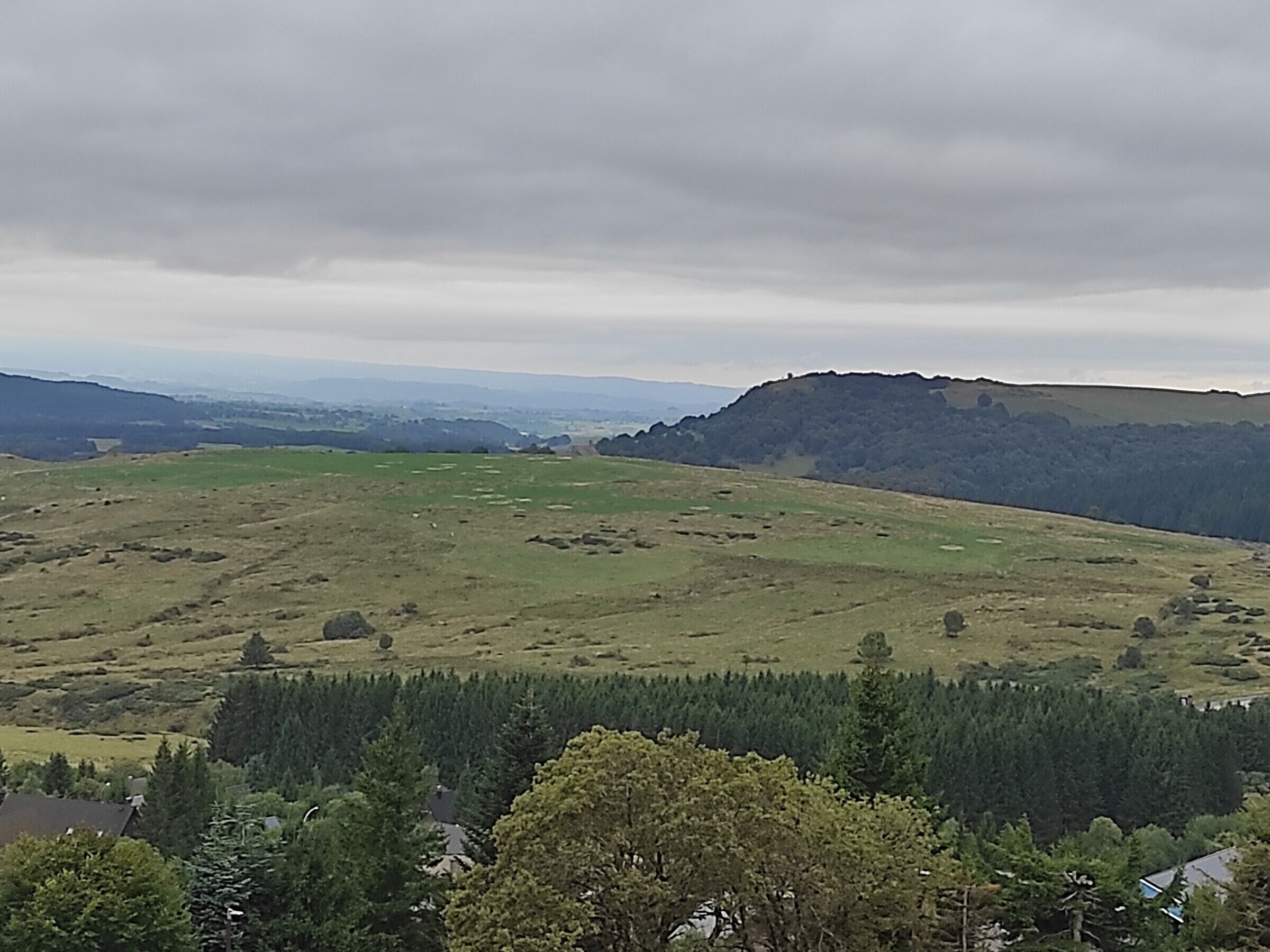 Vista panoràmica d'un paisatge amb zones de muntanya, una àrea boscosa al primer pla i muntanyes llunyanes sota un cel nuvolós.