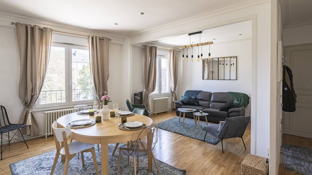 Un espacio habitable amplio con una mesa de comedor dispuesta para cuatro personas, un sofá de color oscuro y un ventanal que proporciona luz natural.
