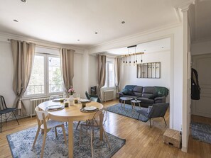 A spacious living area with a dining table set for four, a dark-colored sofa, and a large window providing natural light. - Elegant Duplex in the Heart of Reims - 2 Bedrooms (Reims)